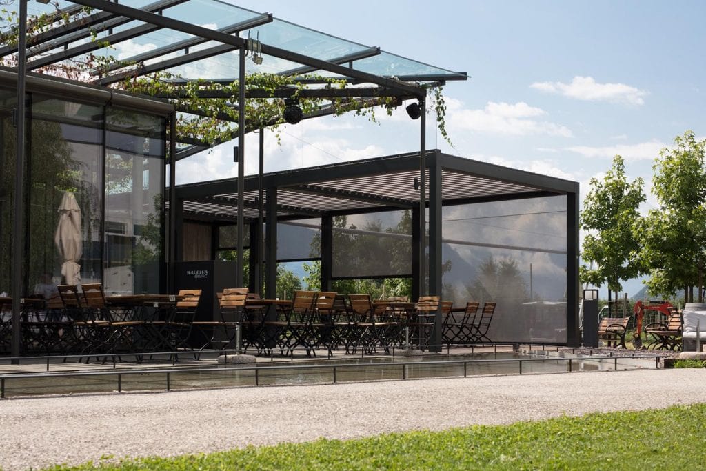 Bioclimatic pergola with adjustable sunscreen blades to protect seats in a bar or restaurant from sun and rain